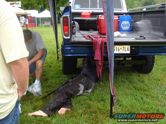 Person working under the rear of a blue pickup truck, with tools and containers in the bed