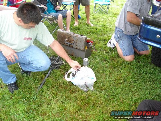 Man in jeans and t-shirt crouches near a toolbox, holding a car part from a plastic bag. Another person kneels by a blue vehicle