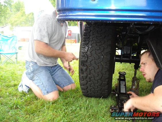 Two guys working on a truck tire with a jack. One is kneeling by the tire, the other is operating the jack