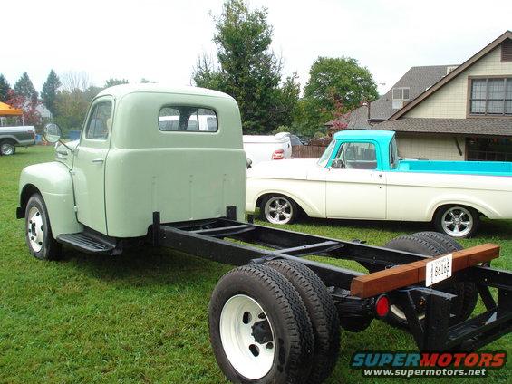 Light green truck chassis with cab, rear view. White truck with blue bed in background
