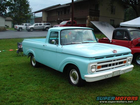 Light blue pickup truck parked on grass, other trucks and building in background