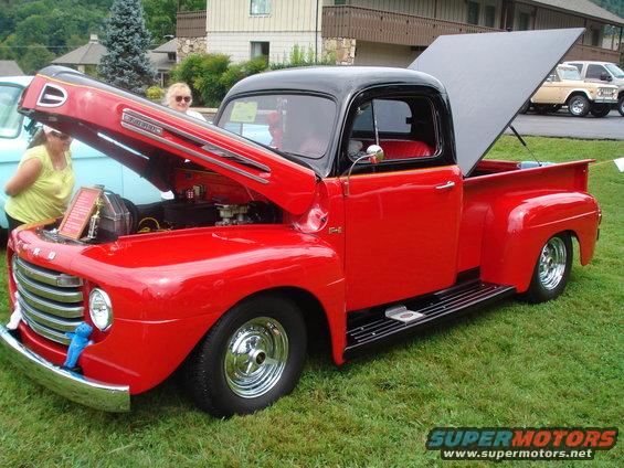 Red 1950s Ford F-1 pickup truck with hood and bed open, parked on grass. Another vehicle is partially visible to the left