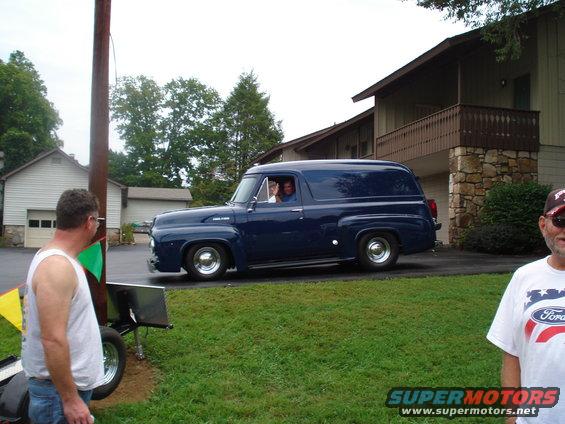 Dark blue 1953 Ford panel van driving past a house, driver waves from window