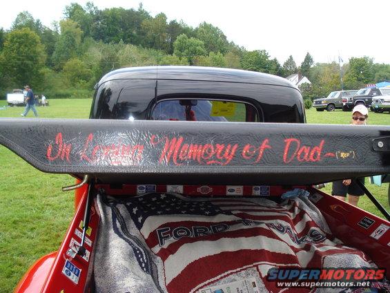Black pickup truck tailgate with red lettering "In Loving Memory of Dad - (RIP)" over an American flag blanket in the bed