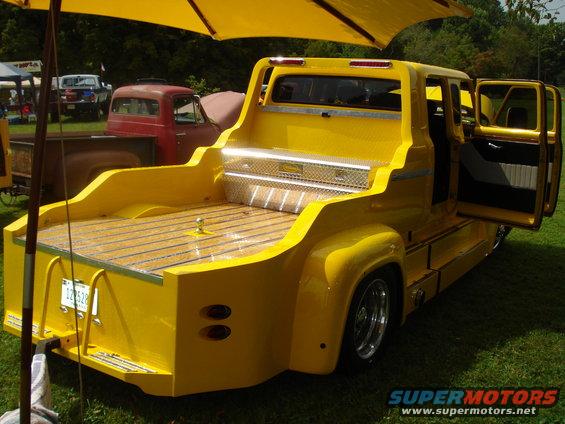 Custom yellow pickup truck with wooden bed and diamond plate toolbox, viewed from rear. Red truck in background