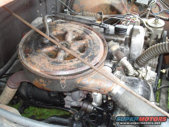 Engine bay with large, rusty air filter housing and visible engine components. A metal rod lies across the housing