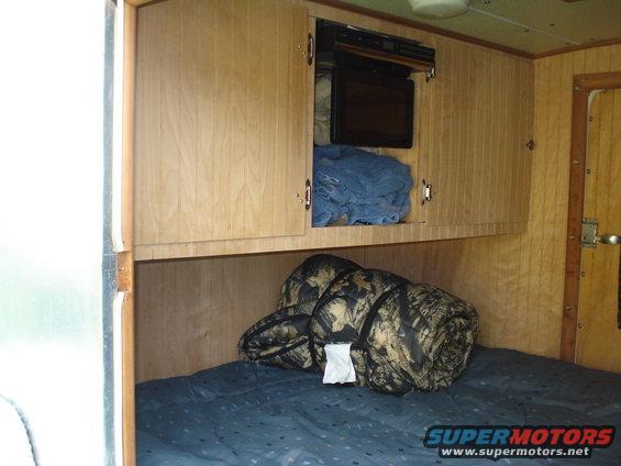 Camper interior with a bed, camouflage sleeping bags, and overhead cabinets with a microwave