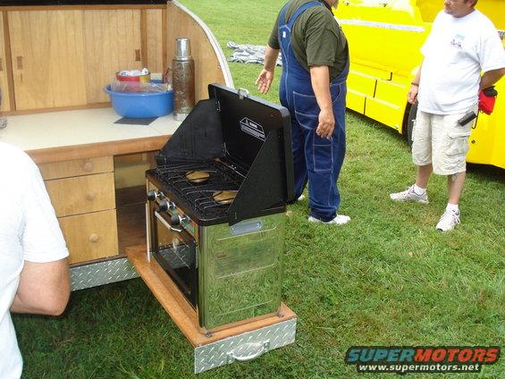 Outdoor kitchen setup with a propane stove and oven, two men standing nearby, and a yellow vehicle in the background