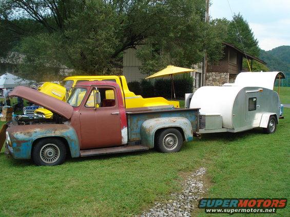 1955 Ford F-100 pickup truck, primer and rust, towing a silver teardrop trailer. Yellow truck behind it