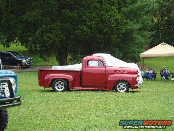 Red classic pickup truck with white cover over bed, parked on grass. Another truck and people in background