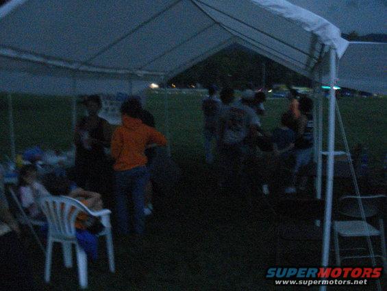 People gathered under a white tent at dusk, with a grassy field in the background