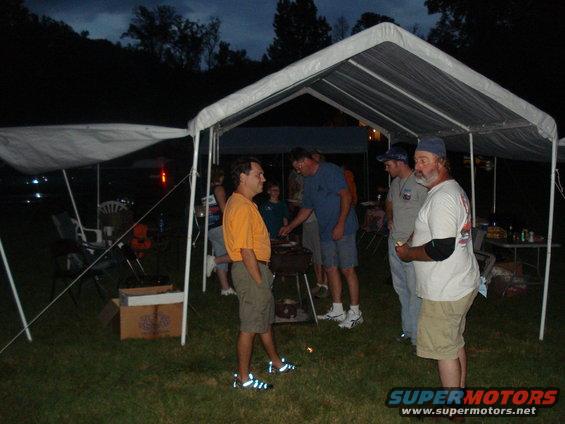 Group of people gathered under white tents at dusk, with a grill visible