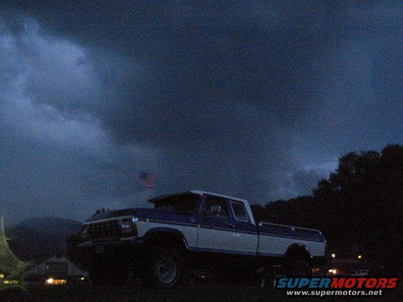 Blue and white pickup truck with an American flag flying under a stormy sky