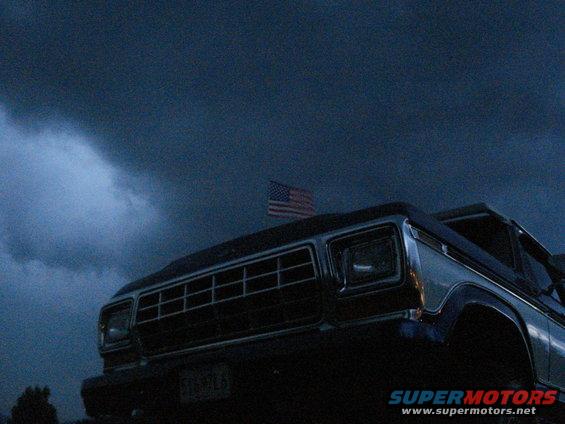 Front view of a dark 1979 Ford F-Series truck with an American flag on top, under a stormy sky