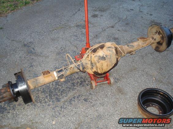 Rear axle assembly with differential housing, brake drum, and wheel studs, supported by red jack stands on asphalt