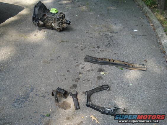 Transmission, control arm, and bracket parts laid out on asphalt
