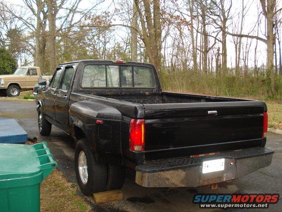 Rear view of a black dually pickup truck parked outdoors, with another older pickup truck visible in the background