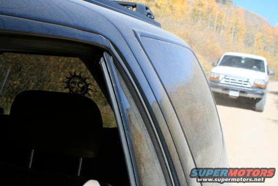 View from inside a dark SUV looking out at a white SUV on a dirt road with autumn trees in background