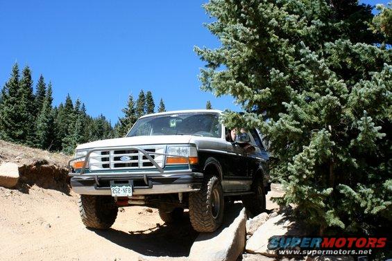 1996 Ford Bronco driving on a rocky, dirt trail with trees on the right