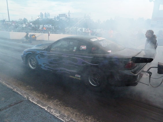 Drag car with purple flames and a large rear spoiler on a race track, emitting smoke
