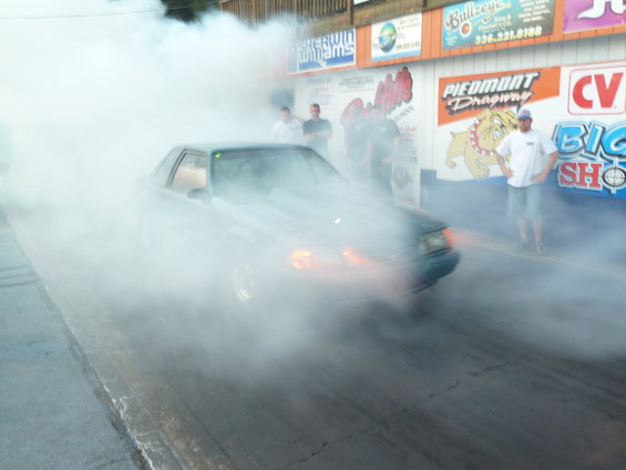 car doing a burnout on a drag strip, kicking up smoke. Signs on the wall behind it