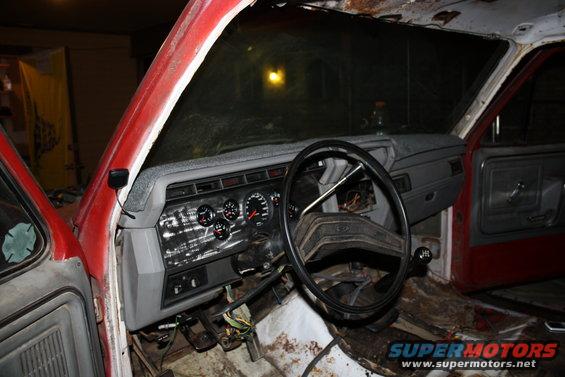 Red truck interior with worn dashboard, steering wheel, and floor. Aftermarket gauges are visible