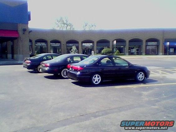 Three dark sedans parked in a lot, one in the foreground with a red taillight strip
