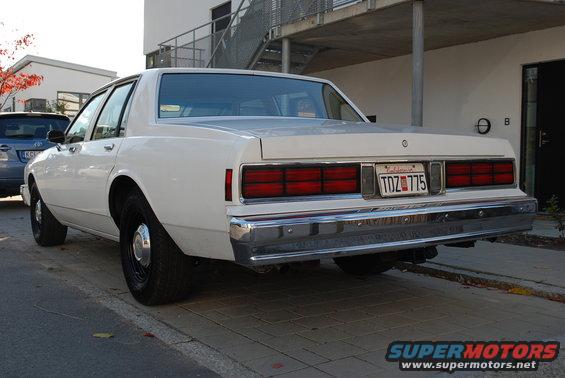Rear view of a white 1980s sedan parked on a sidewalk. It has a chrome bumper and horizontal taillights