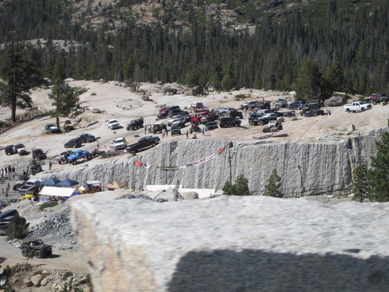 Many off-road vehicles parked on a rocky, wooded hillside near a quarry wall with banners