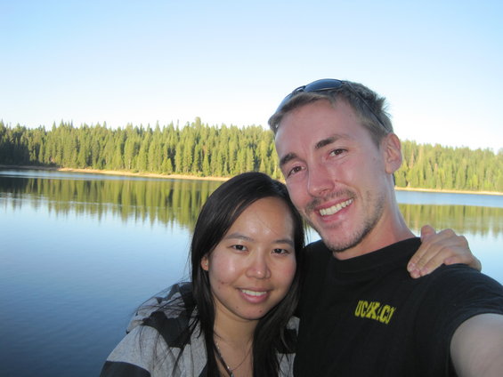 Couple smiling for a selfie in front of a lake with a dense forest in the background