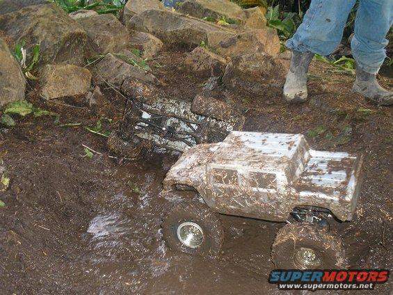 Two mud-covered remote control trucks stuck in mud and grass with rocks in the background and a person's legs in jeans and boots standing nearby
