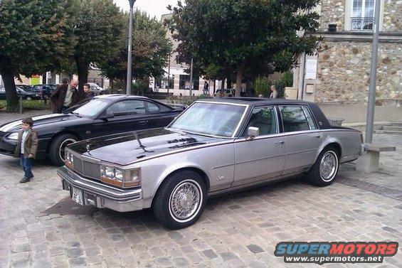 Silver 1980s Cadillac Seville with black hood and wire wheels parked next to a black Ford Mustang