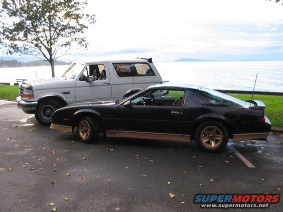 Side view of a white Ford Bronco parked next to a black Chevrolet Camaro by the water
