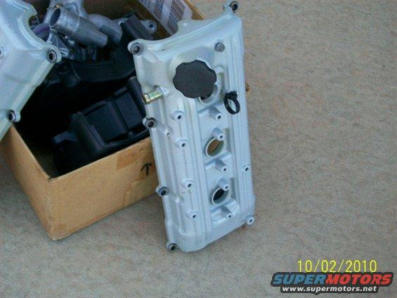Silver valve cover with black cap and ring, in a cardboard box with other engine parts