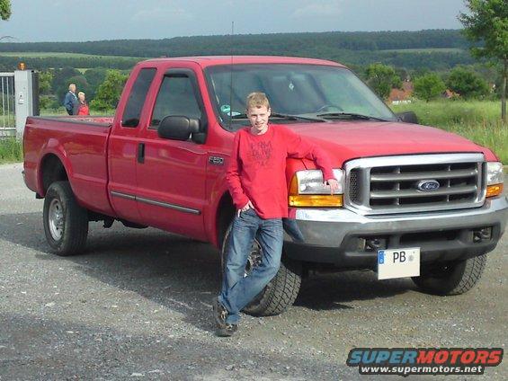 Red Ford F-250 pickup truck with a young man leaning on the hood. The truck has a crew cab and a long bed
