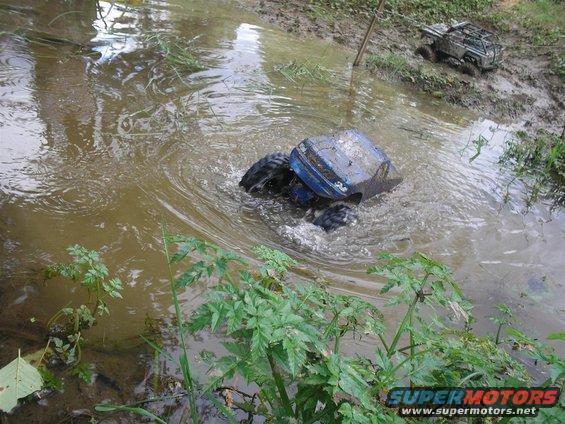 Blue remote control car partially submerged in muddy water, with another RC vehicle in the background
