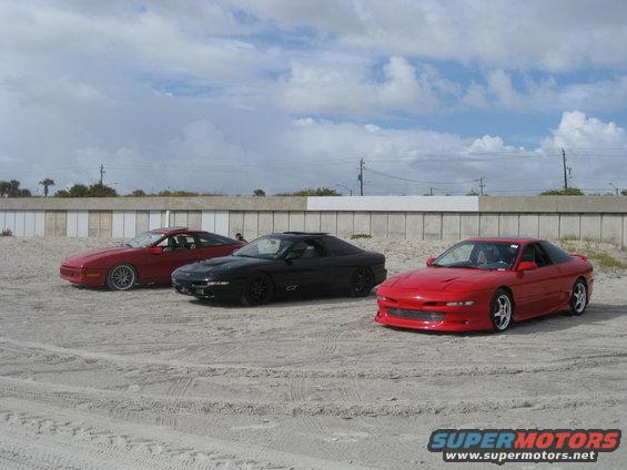 Three 1994 Ford Probes parked on a sandy area with a wall and cloudy sky in the background
