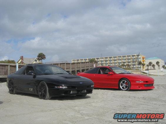 Black and red 1994 Ford Probe cars parked on a beach with buildings and palm trees in the background