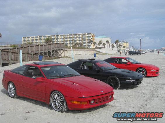 Three Ford Probe cars parked on a sandy beach with a hotel in the background