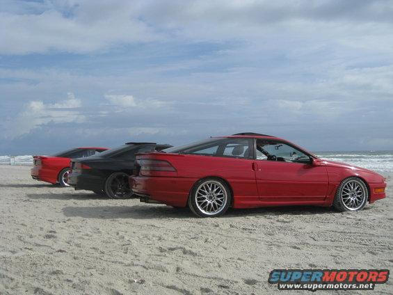 Red coupe with aftermarket wheels parked on a sandy beach next to two other coupes