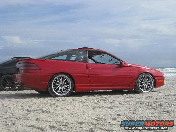 Red 1994 Ford Probe coupe parked on a sandy beach with the ocean in the background