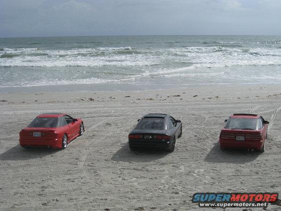 Three coupes parked on a sandy beach facing the ocean; two red, one black
