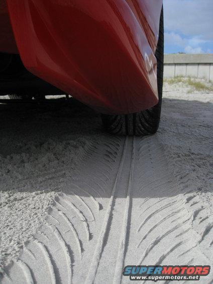 Tire tracks left by a red car on a sandy surface near a concrete barrier
