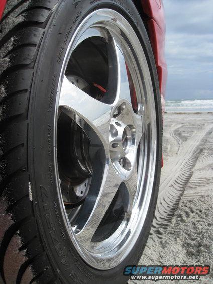 Red car's chrome five-spoke wheel and tire on sandy beach with tire tracks and ocean in background