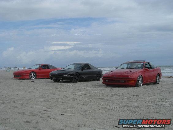 Three red and black Ford Probe cars parked on a sandy beach with the ocean in the background