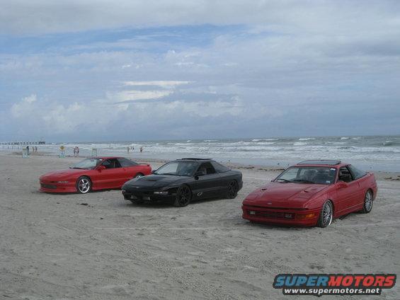 Three red and black 1994 Ford Probes parked on a sandy beach with the ocean in the background