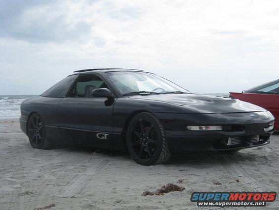 Black Ford Probe GT parked on a sandy beach with the ocean in the background. Another red car is partially visible to the right