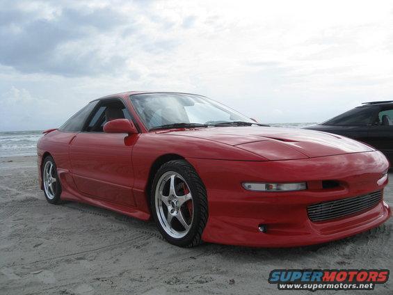 Red sports coupe parked on a sandy beach with the ocean in the background. It has aftermarket wheels and a custom grille