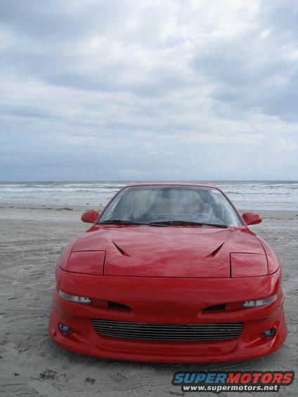 Red sports car with pop-up headlights parked on a sandy beach with the ocean in the background