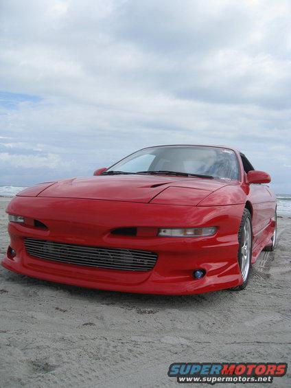 Red sports car parked on a sandy beach with the ocean and cloudy sky in the background. It has a custom grille and body kit